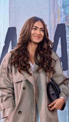 a woman standing in front of a wall holding a black purse and smiling at the camera