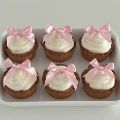 six cupcakes with white frosting and pink bows in a baking pan on a table