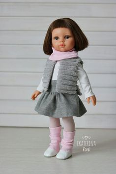 a doll with brown hair wearing a gray dress and pink scarf is standing in front of a white wall