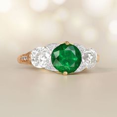 A Stunning Three Stone Ring That Features A Round Cut Natural Emerald Luxury Emerald Birthstone Ring With Center Stone, Fine Jewelry Emerald Three-stone Diamond Ring, Emerald Three Stone Diamond Ring In Fine Jewelry Style, Green Three Stone Diamond Ring, Luxury Green Three Stone Diamond Ring, Timeless Emerald Ring With Three Stones, Luxury Green Diamond Three Stone Ring, Luxury Green Three-stone Diamond Ring, Luxury Three-stone Green Diamond Ring