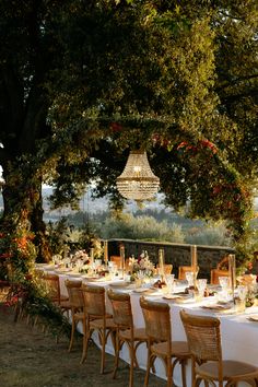 an outdoor dining table set up with white linens and wooden chairs, surrounded by greenery