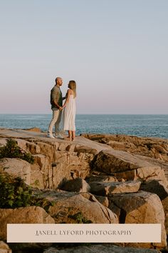 a man and woman standing on rocks near the ocean with their arms around each other