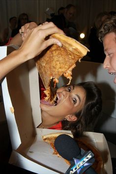 a woman is eating pizza from a box while someone holds her hand over the mouth