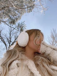 a woman with long hair wearing ear muffs and fur coat in front of trees