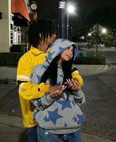 a man and woman standing next to each other on a sidewalk at night with street lights in the background