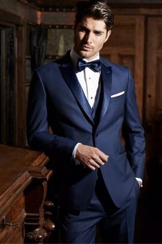 a man in a blue tuxedo standing next to a wooden table wearing a white shirt and bow tie