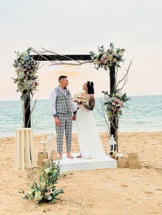 a man and woman standing on top of a sandy beach next to an arch covered in flowers