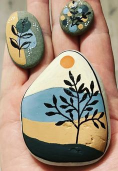 two painted rocks in the palm of someone's hand, one with a tree on it