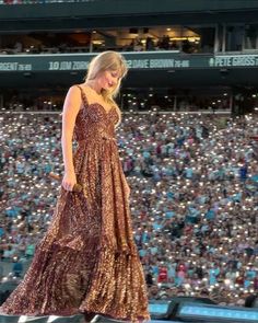 a woman in a long dress walking across a stage with an audience behind her at a baseball game