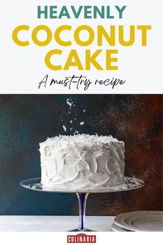 Tall layer cake with swirled white frosting and fresh coconut shavings sprinkled on top sits elegantly on a blue-stemmed cake stand against a moody dark background.