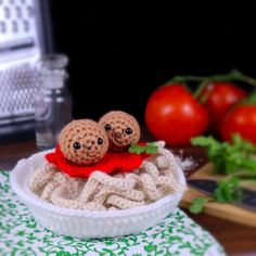 two tiny crocheted dolls sit in a bowl on a table next to tomatoes