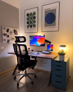 a desk with two monitors and a lamp on it in front of some pictures hanging on the wall