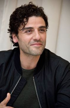 a man with curly hair and a black jacket giving the thumbs up while sitting in front of a white curtain