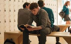 a man sitting on a bench reading a book while others look at lockers in the background