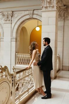 a man and woman standing next to each other in front of a balcony with gold railings