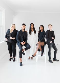 five people posing for a photo in front of a white wall and window, with one person sitting on a chair