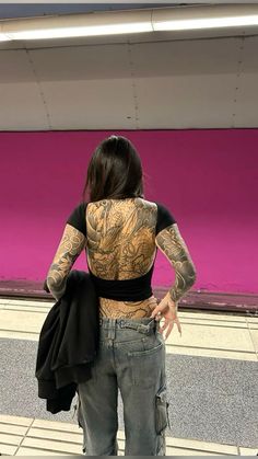 a woman with tattoos on her back standing in front of a pink wall and looking at the ground