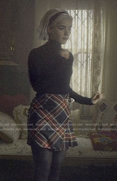 a woman standing in front of a window wearing a plaid skirt and black top with her hands on her hips