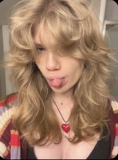 a woman sticking her tongue out in front of the camera with long blonde hair, wearing a red heart necklace