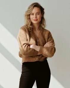 a woman standing with her arms crossed and looking at the camera while wearing a brown cardigan