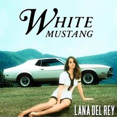 a woman sitting on the ground in front of a white mustang with mountains in the background