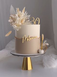 a white cake with gold lettering and flowers on top is sitting on a stand in front of a gray backdrop