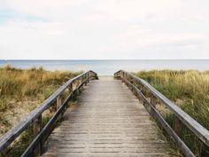 a wooden walkway leading to the beach with tall grass and water in the background on a cloudy day