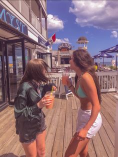 two young women standing on a wooden deck talking to each other while holding drinks in their hands