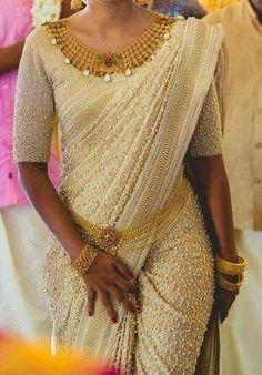 a woman in a white sari with gold jewelry on her neck and hands behind her back