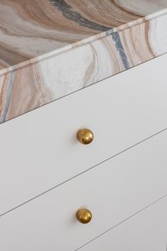 a white dresser with two gold handles and marble counter top on the bottom, in front of a wall