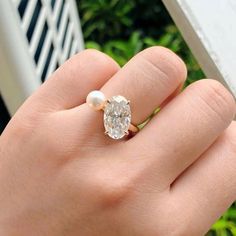 a person's hand holding a ring with a diamond and pearl on it, in front of some bushes