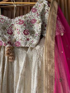 A three-piece ivory and fuchsia bridal lehenga set from the Priti Sahni collection. This beautiful georgette lucknowi thread work lehenga with a heavy border of gota, pearl, sequin, zari work border in pink accent detail is paired with a raw silk blouse with mirror, thread zardozi work all over it. The lehenga has side hanging ball tassels to the waistline. And the blouse has a sequins tassel tie-up at the back. This outfit is completed with a fuchsia net mirror butti dupatta with zari work bord Designer Raw Silk Lehenga With Chikankari Embroidery, Designer White Dola Silk Lehenga, White Dola Silk Lehenga For Designer Wear, Designer Off-white Lehenga With Pallu, White Choli With Intricate Embroidery In Raw Silk, Cream Zari Work Georgette Choli, White Raw Silk Lehenga With Intricate Embroidery, White Lehenga With Intricate Embroidery In Raw Silk, Cream Choli With Zari Work In Georgette