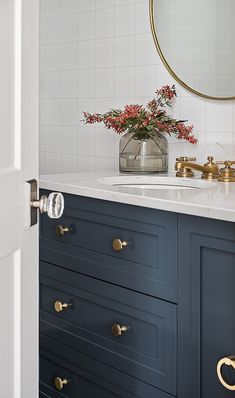 a bathroom with blue cabinets and white tile on the walls, gold faucets and a round mirror