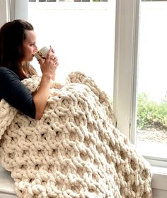 a woman holding a coffee cup while wrapped up in a large chunky knit blanket