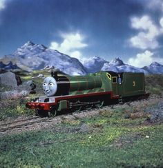 thomas the tank engine is coming down the tracks in front of some mountains and clouds