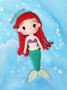 a crocheted mermaid doll with red hair and green tail sitting on blue fabric