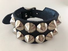 "Classic studded punk bracelet with 2 rows of 1/2\" cone studs. Fastens with a buckle and is adjustable. All hardware is nickel plated. Studs on black bracelets are nickel plated brass, studs on colored bracelets are nickel plated steel (old stock) Smaller bracelets have fewer studs per row. Made with vegetable tan leather. Note: red bracelet is kind of pink-ish. Bracelet is 1.25\" wide. Ask about custom sizing and color options!" Punk Black Bracelet With Silver Studs, Silver Rivets Wristband For Concert, Rocker Style Leather Bracelet With Studs For Festivals, Adjustable Silver Bracelet With Spikes, Trendy Adjustable Spiked Bracelets, Metal Studs Bracelet For Concerts, Metal Stud Bracelets For Concerts, Adjustable Punk Cuff Bracelet With Rivets, Metal Studded Bracelets For Concerts