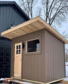 a small shed sitting in the snow next to two garage doors and a building with windows