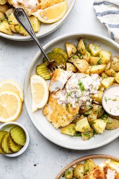 two plates with chicken, potatoes and pickles on them next to lemon wedges