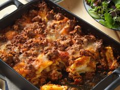 a casserole dish with meat and cheese in it sitting on a table next to a salad