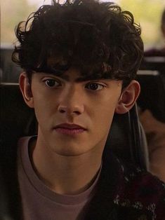 a young man with curly hair sitting in a car looking at the camera while another person sits behind him