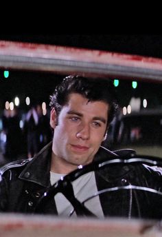 a man in a black leather jacket is looking at the camera while standing next to a car