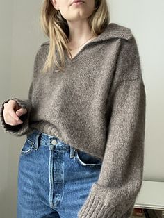 a woman standing in front of a white wall wearing a gray sweater and blue jeans