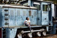 a woman sitting on the side of a blue train next to some trees and bushes