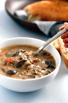 a person holding a spoon in a bowl of soup next to some bread on a plate