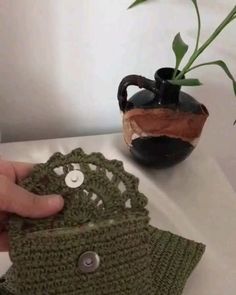 a hand is holding a green crocheted pot holder next to a small plant