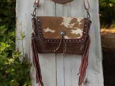 Such a beauty, stamped genuine chocolate brown leather on the bottom, with a flap closure that features hair on hide, leather stitching accents around the sides, and a concho with leather tie accents. The sides are adorned with long brown fringe, and the back features the same hair on hide on the top section, and stamped leather on the bottom.  It even includes a zippered section for each access to all your necessities. Myra bags are so unique, each one crafted from genuine leather, some with one of a kind hair on hide, or canvas crafted from recycled military tarps.  The leather straps are totally adjustable, use it as a shoulder purse, or lengthen the strap and use it as a crossbody. The bag's cotton lining is printed with the "Myra" logo, and most offer inside open storage pockets and a Western Style Brown Bag With Concho, Western Brown Bag With Adjustable Strap, Western Brown Shoulder Bag With Adjustable Strap, Western Style Brown Shoulder Bag With Adjustable Strap, Purse With Fringe, Myra Bags, Cowhide Purse, Brown Fringe, Stamped Leather