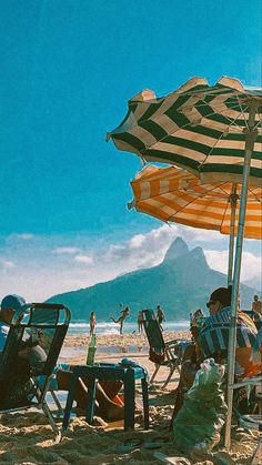 people are sitting under umbrellas on the beach near the water and mountains in the background