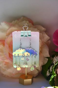 a pair of earrings is hanging from a stand with flowers in the back ground and a pink flower behind it