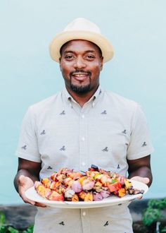 a man in a hat holding a plate with food on it and smiling at the camera